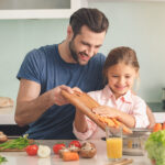 Father and a daughter standing in a kitchen putting chopped carrots into a pot.